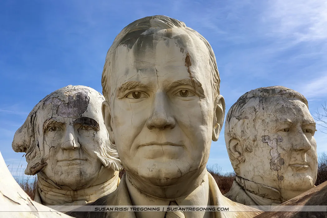 Large, weathered concrete busts of Calvin Coolidge, Martin Van Buren, and Millard Fillmore stand outdoors beneath a clear blue sky. The sculptures show cracks, discoloration, and surface erosion from long exposure to the elements.