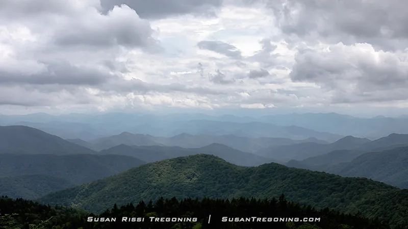 Layered blue mountain ridges stretch into the distance at Cowee Mountain Overlook as storm clouds move in. Dense green forest fills the foreground, and the fading ridgelines recede into a hazy, overcast sky.