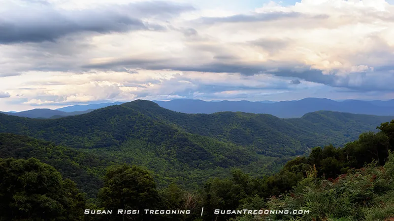A wide mountain panorama is seen from Lane Pinnacle Overlook, with layers of blue ridges fading into the distance under early‑evening light.