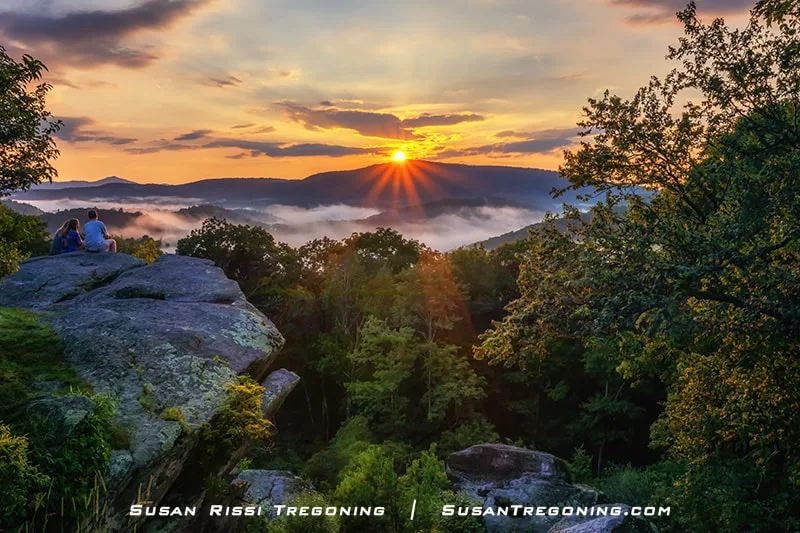 A sunset casts warm light across the Blue Ridge Mountains from Raven Rocks Overlook at milepost 289.5. Two people sit on a large rock formation in the foreground, looking out over a mist‑filled valley and layers of forested ridges. The sky glows with orange and yellow tones above the distant mountains.