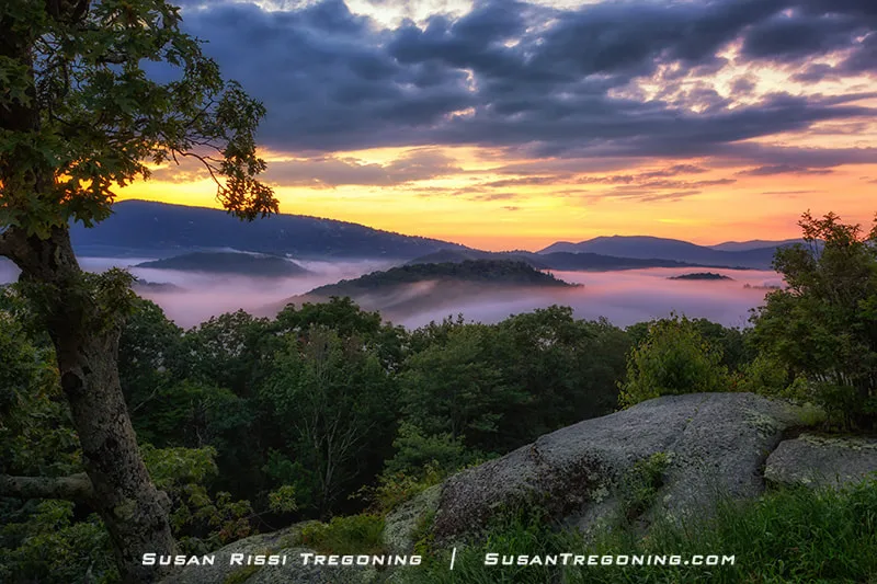 A sunset glows over the Blue Ridge Mountains from Raven Rocks Overlook at milepost 289.5. Clouds settle in the valleys, creating the appearance of small mountain islands rising through the mist. Warm light reflects across the tops of the clouds, with layered ridge lines stretching into the distance.