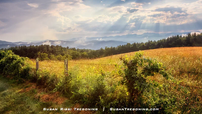 A pastoral landscape shows a grassy field bordered by a wire fence and dense green vegetation, with forested hills and distant Blue Ridge Mountains in the background. Sunlight breaks through the clouds, creating bright rays that shine down across the scene.