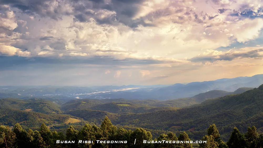   
A wide mountain landscape shows layers of green, forested ridges stretching into the distance under a sky filled with soft, glowing clouds. Sunlight filters through the clouds, creating warm highlights across the hills and valleys.