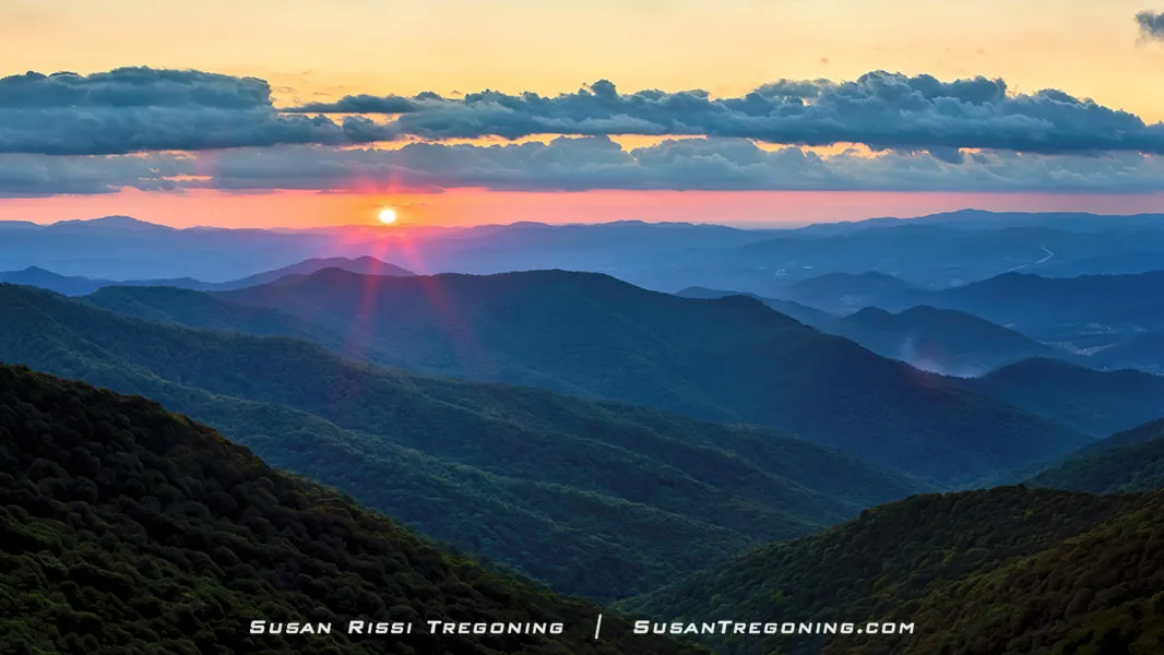 The sun sets over the Blue Ridge Mountains near Craggy Gardens, casting warm orange and pink light across the sky. Layered mountain ridges fade into the distance in deep blue tones, with forested slopes in the foreground. Thin clouds catch the sunlight near the horizon, creating a soft glow above the peaks.
