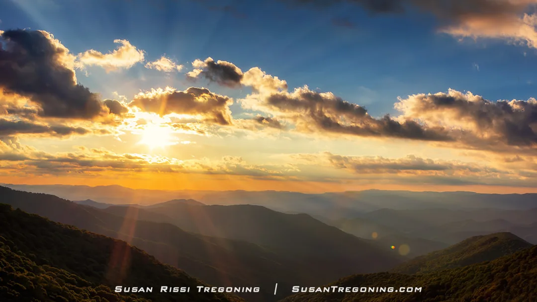 The sun sits low in the sky above the Blue Ridge Mountains near Craggy Gardens, with bright golden light radiating outward in multiple directions. Sunbeams stream through breaks in the clouds, creating a dramatic ring of rays across the sky. Layered mountain ridges fade into the distance, shifting from warm tones near the horizon to cooler blue hues farther away.