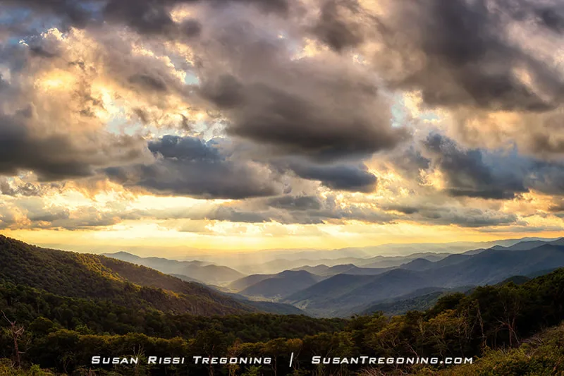   
Sunlight breaks through thick clouds over the Blue Ridge Mountains near Glassmine Falls Overlook. Rays of warm golden light stream down into the valley, illuminating layered ridgelines and forested slopes. Darker clouds hang above the scene, creating a dramatic contrast between shadowed peaks and the sunlit landscape. The overlook itself is not visible in the frame.