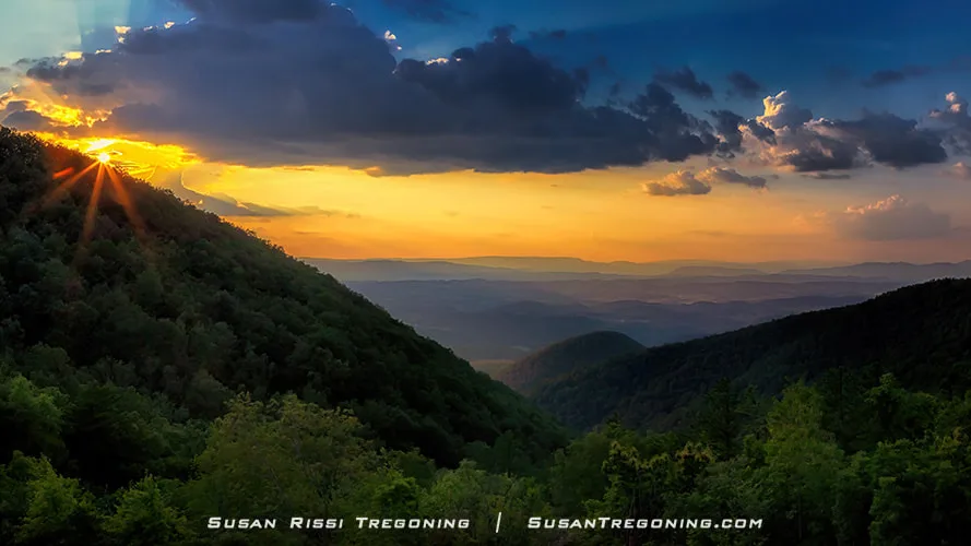  The sun sets behind a mountain ridge at Taylors Mountain Overlook, with warm rays shining through breaks in the clouds. Forested hills fill the foreground, and distant blue mountain layers fade toward the horizon beneath a partly cloudy sky.