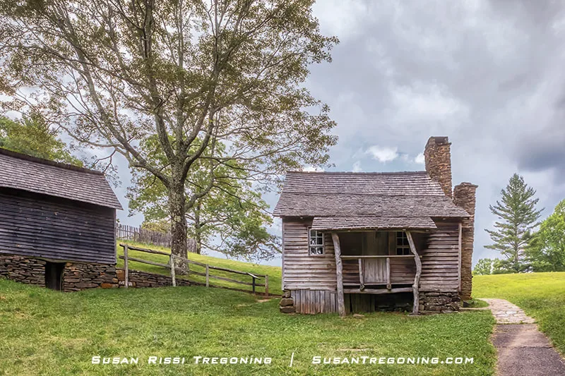 A one‑story log cabin with lapped wood siding sits on a stacked fieldstone foundation at Brinegar Cabin. A crooked wooden post supports the front porch roof, and the cabin is surrounded by grass and trees.