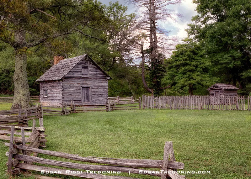 A small log cabin sits in a grassy clearing on Groundhog Mountain, surrounded by trees. The simple wooden structure has a stone chimney on one end and a pitched roof, reflecting its historic Appalachian construction.