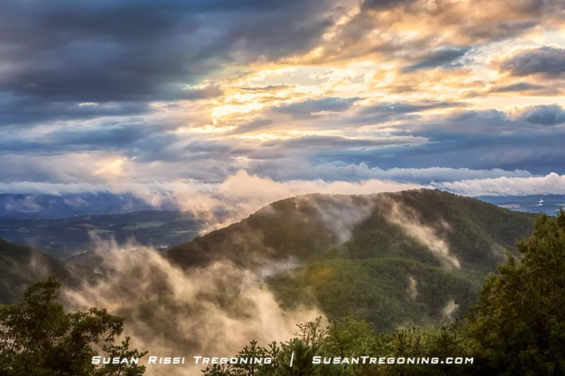Low‑lying clouds drift across the mountains at Irish Creek Overlook, partially obscuring the normally wide view. Mist clings to the green ridges, and dramatic clouds above catch warm light from the rising or setting sun. Leafy vegetation frames the foreground.