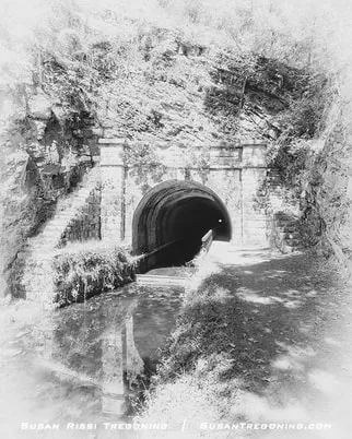 The brick entrance of the Paw Paw Tunnel sits at the base of a rocky, tree‑covered hillside, with a narrow canal leading directly into the dark arched opening. Vegetation lines the path beside the water, and the scene is softly lit.