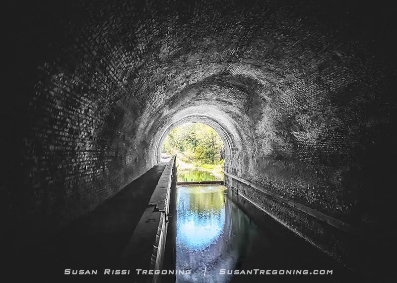 Inside the Paw Paw Tunnel, dim brick walls curve overhead as still canal water reflects the bright light and greenery visible at the far opening.