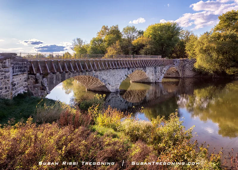 A historic stone arch aqueduct spans a calm, tree‑lined river, its multiple arches and wooden railings reflected in the water below. Warm late‑day sunlight illuminates the stonework and surrounding greenery.