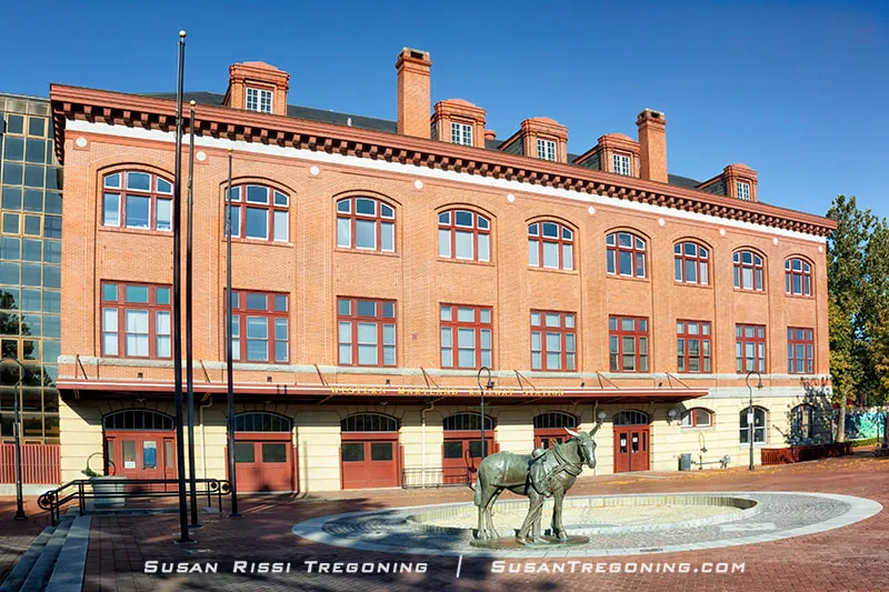 The Western Maryland Railway Station, a large three‑story brick building with rows of arched windows, stands behind a circular fountain featuring a bronze mule‑and‑boat statue, all set within a brick plaza under a clear blue sky.