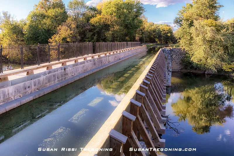 A view looking down the historic Conococheague Aqueduct shows calm canal water flowing between stone walls with wooden railings, a pedestrian walkway running alongside, and lush green trees surrounding the structure.