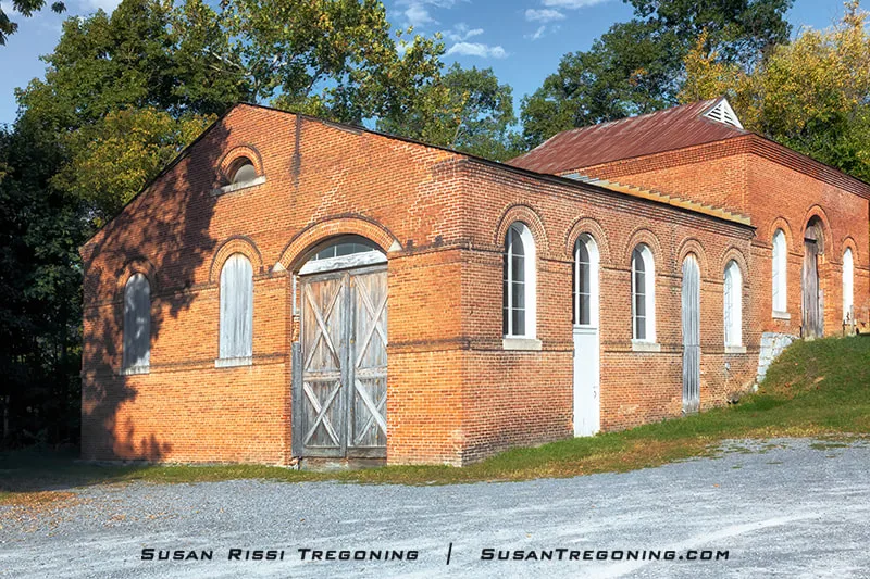 A historic red‑brick trolley barn with arched windows and large wooden double doors stands beside Cushwa’s Warehouse in a grassy, tree‑lined setting. A gravel drive leads to the building, and a small vented cupola sits on the metal roof.