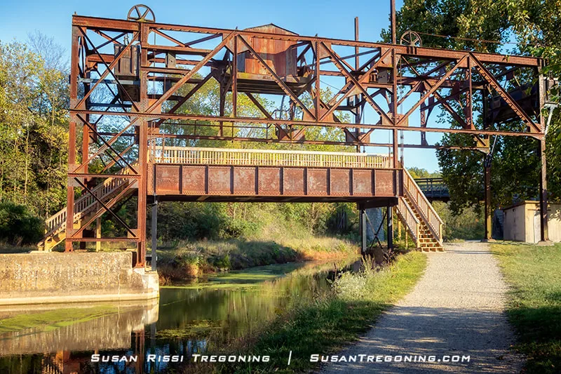 A historic steel lift bridge spans a narrow canal, its rusted truss frame rising above the water with staircases on both sides leading to a central walkway. Gears and pulleys sit at the top of the structure, and the canal is surrounded by grassy banks, trees, and a gravel path.