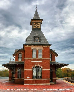 A historic red‑brick train station with a steep slate roof and a tall central tower topped by a spire stands beside a brick platform. The tower features a circular decorative window and arched openings, and autumn trees surround the building under an overcast sky.