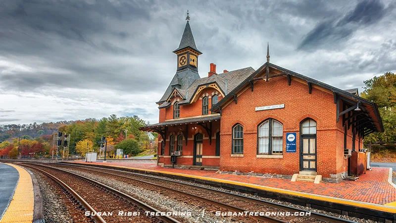 A historic brick train station with a steep roof, decorative trim, and a clock tower stands beside railroad tracks lined with yellow tactile paving. Autumn trees in shades of green, orange, and red surround the building under a cloudy sky.