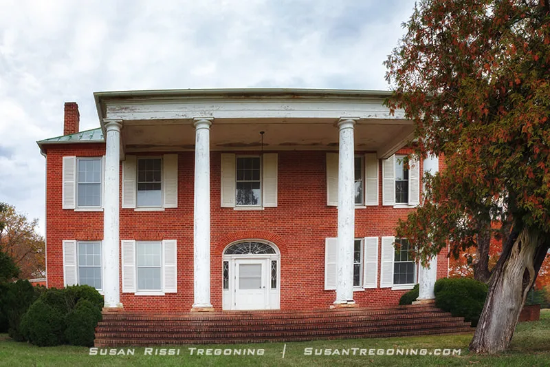 A large red‑brick, two‑story historic house features a columned white portico with four tall columns, symmetrical windows with white shutters, and brick steps leading to the front door. Trees and shrubs surround the home beneath a partly cloudy sky.