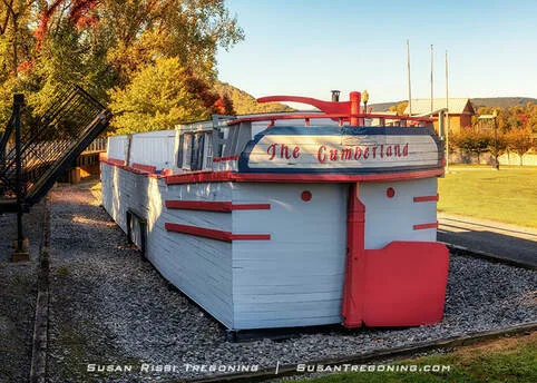 A full‑scale white canal boat replica with red trim, named The Cumberland, sits on a gravel display pad surrounded by grass, autumn trees, and nearby buildings.