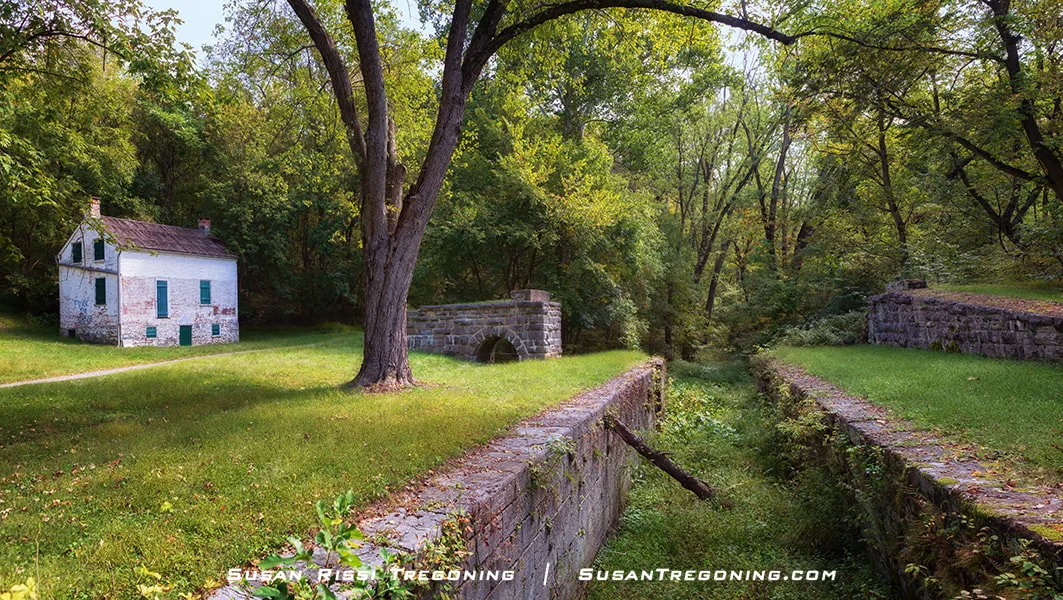 A historic stone lock runs through a wooded clearing, bordered by grassy embankments and dense green trees. A small white brick lockhouse with green shutters and a red roof stands beside a dirt path to the left, with a large tree rising near the lock.
