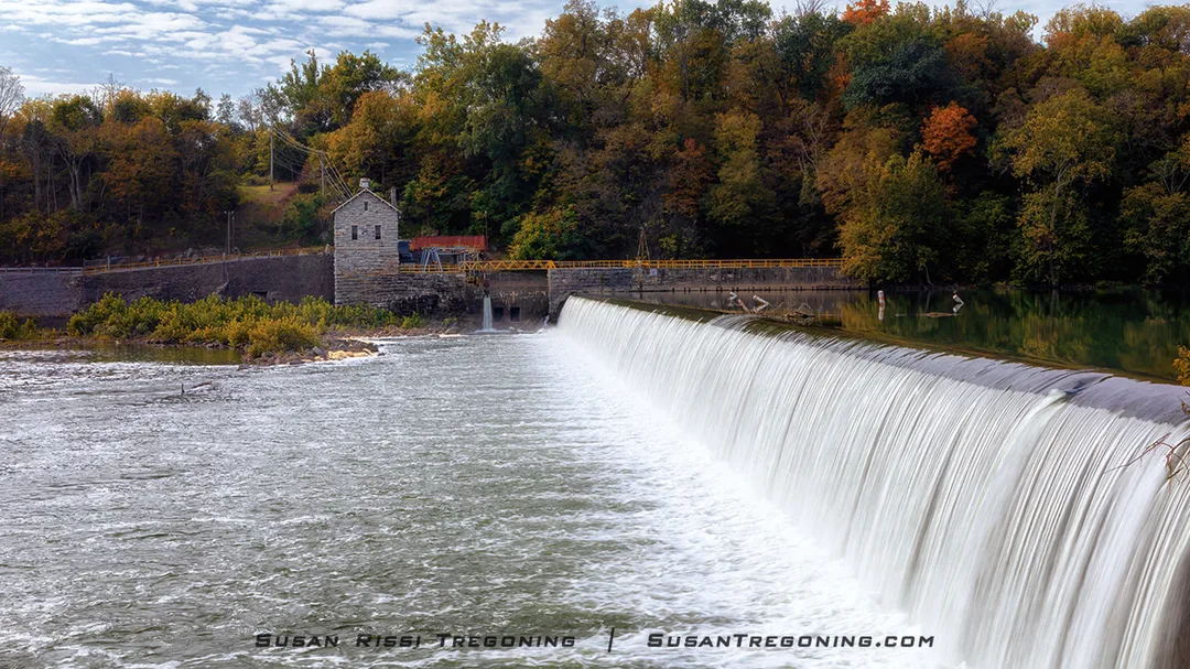 Water cascades evenly over a wide dam, creating a smooth waterfall effect. A small stone building with a pitched roof stands just above the dam, surrounded by autumn trees in shades of green, yellow, and orange beneath a partly cloudy sky.