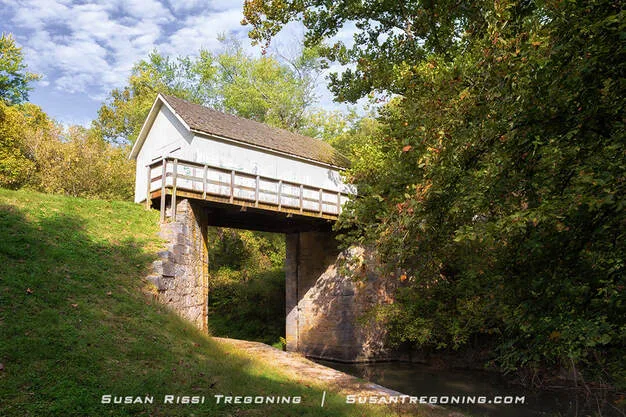A small white wooden building with a shingled roof stands beside a narrow creek, supported by stone abutments. Green trees and grass surround the structure on a sunny day with scattered clouds.
