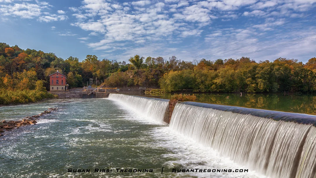 A smooth sheet of water cascades over Dam 5 into the river below, with a red‑brick mill building set among autumn trees in the background under a partly cloudy sky.