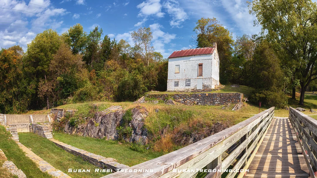 A small historic stone lockhouse with a red roof stands on a raised stone foundation beside the Guard Lock, reached by a wooden footbridge and surrounded by lush green trees under a partly cloudy sky.