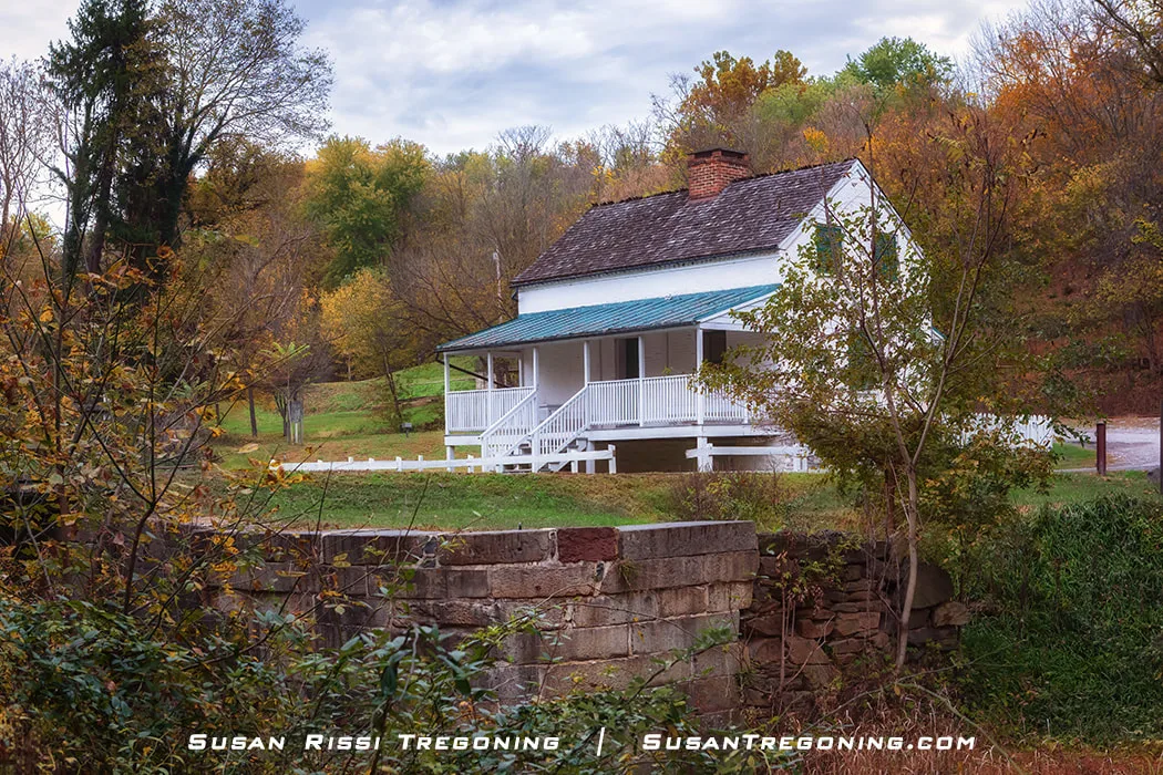 A white historic house with a green metal roof and a brick chimney sits among autumn trees in shades of green, yellow, and brown. A front porch with white railings faces a grassy yard, and an old stone structure lies in the foreground, partially covered by vegetation.