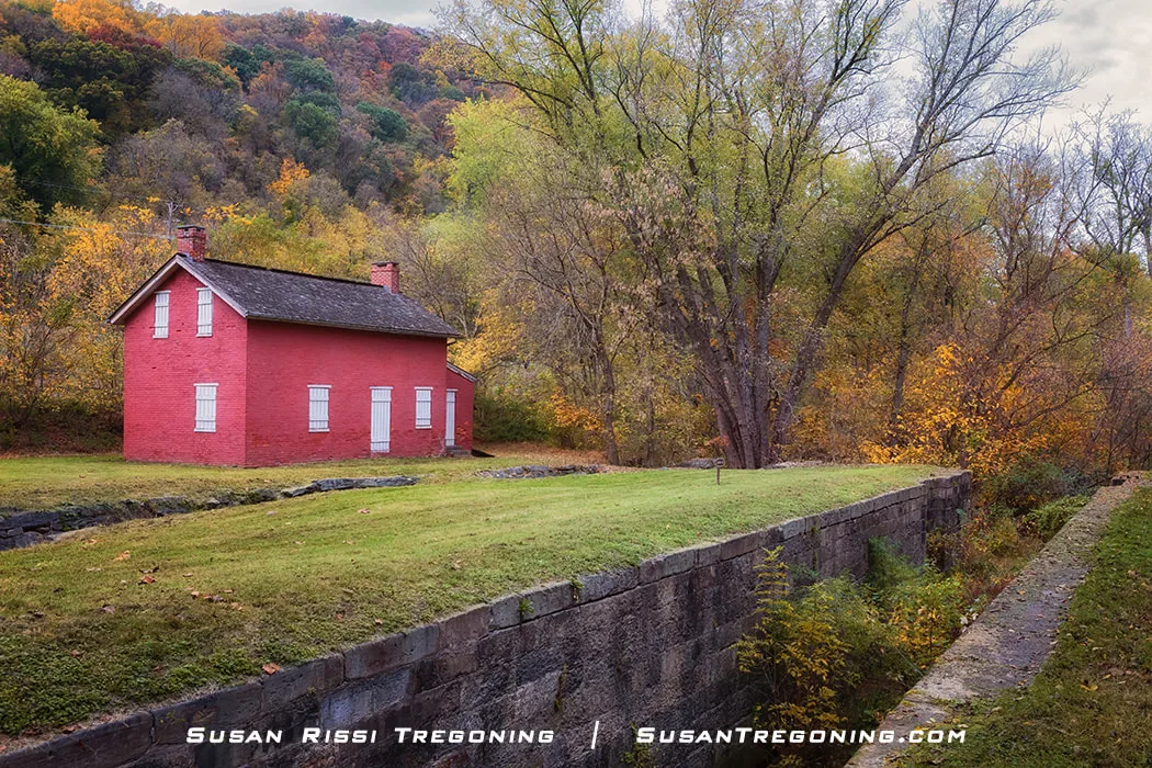 A small red‑brick lockhouse with white shutters stands beside a grassy canal bed and a low stone wall, surrounded by autumn trees in shades of green, yellow, and orange. Forested hills rise in the background, creating a peaceful fall landscape.
