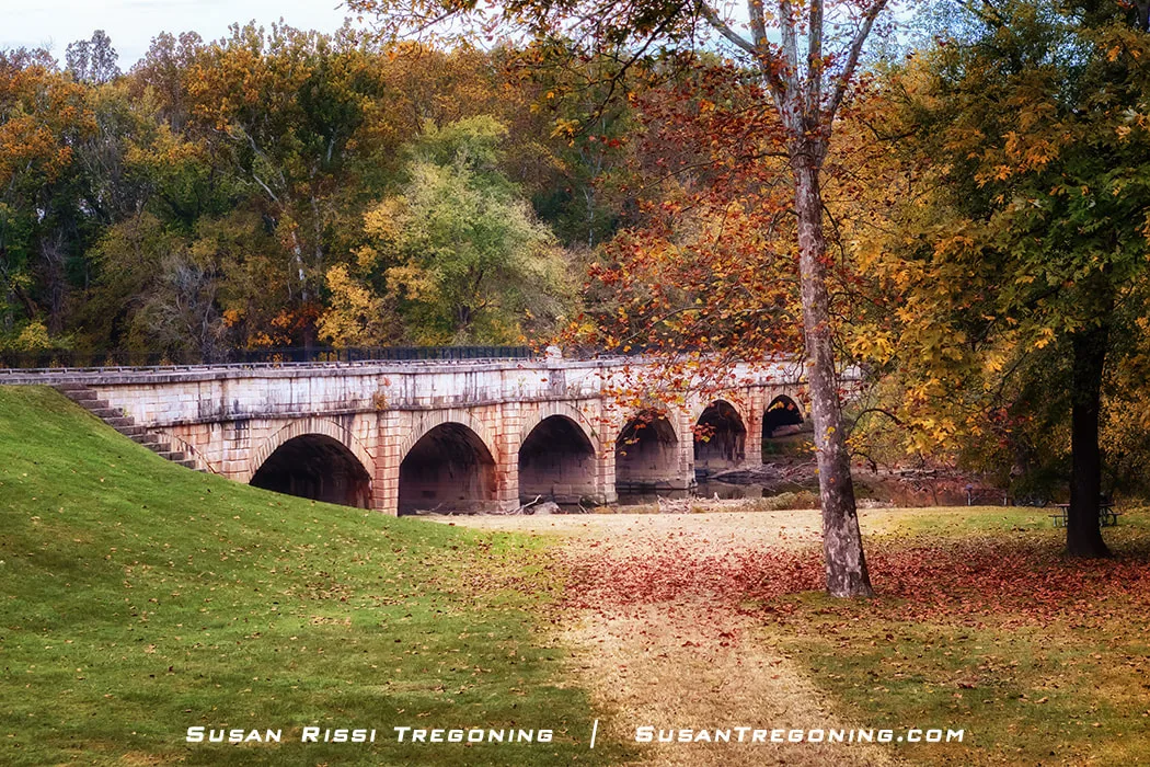 A multi‑arch stone aqueduct carries the historic canal over a shallow stream, surrounded by autumn trees in shades of green, yellow, and orange. Fallen leaves cover the grassy foreground, creating a quiet, picturesque fall scene.