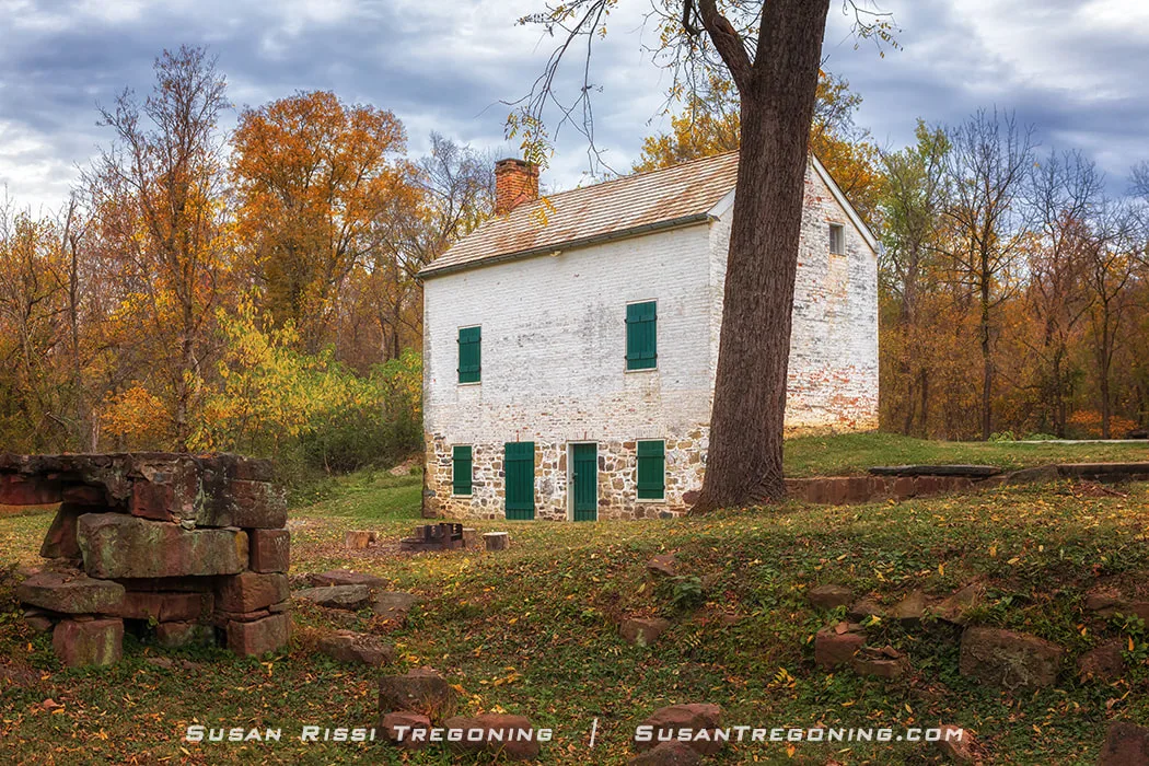 A whitewashed two‑story lockhouse with green shutters stands beside stacked stone ruins and autumn trees in shades of orange, yellow, and brown. Fallen leaves cover the grassy ground, and the scene has a quiet, historic, rural atmosphere.