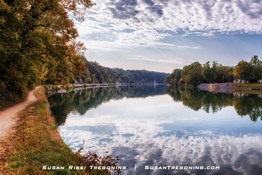 A wide, calm stretch of the Potomac River forms a lake‑like expanse known as Big Slackwater, reflecting the sky and clouds. Dense autumn trees line both sides of the river, with a dirt towpath running along the left bank and a grassy right bank with a few small structures. The still water and soft light create a peaceful, symmetrical scene.