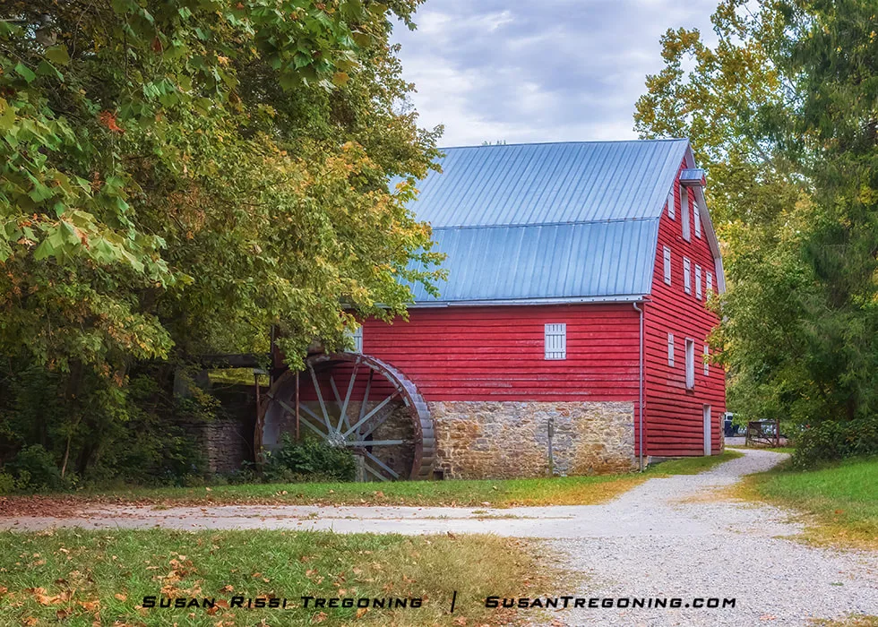 A large red wooden mill with a gray metal roof stands on a stone foundation beside a gravel path, with a tall water wheel attached to one side. Green trees and dense vegetation surround the historic structure, creating a quiet rural scene.