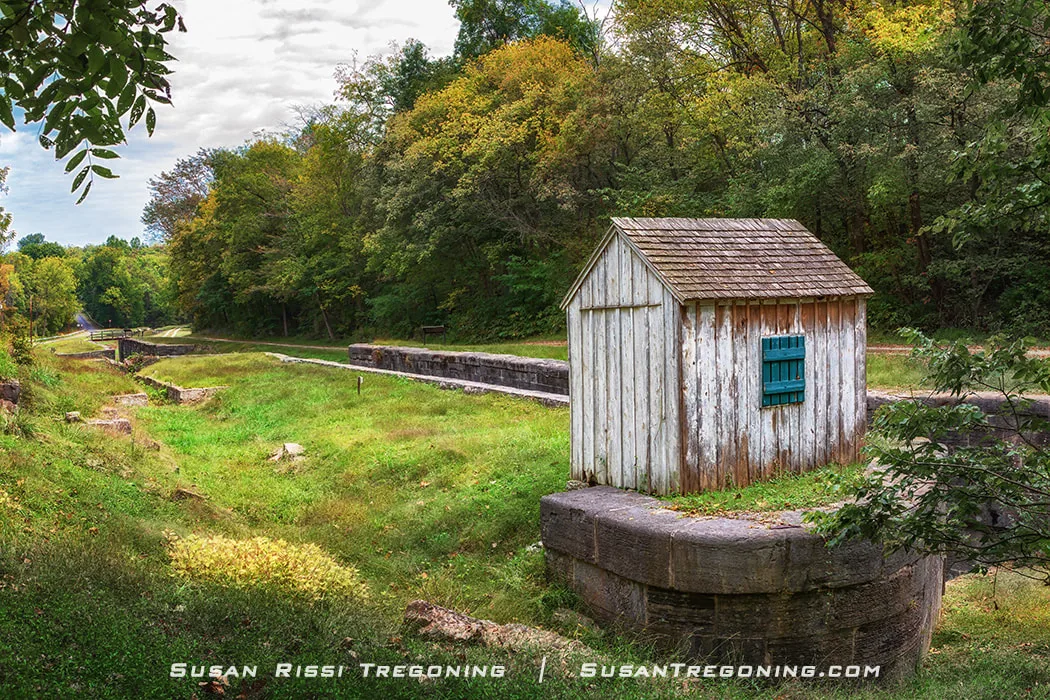 A small weathered wooden Watch House with a shingled roof and blue‑green shutter stands on a stone foundation beside a dry stone canal lock, surrounded by dense trees with early autumn foliage.