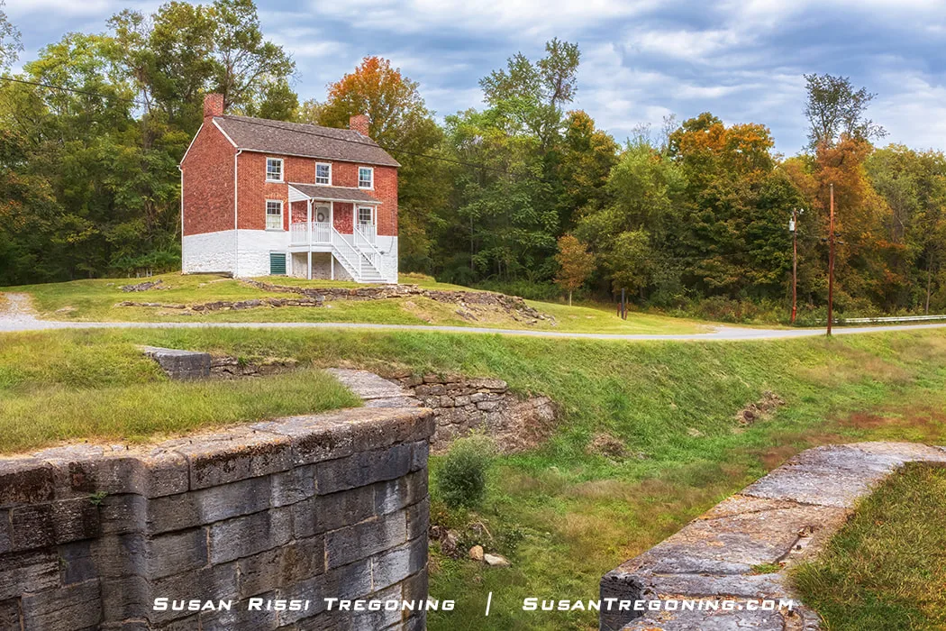 A historic white brick lockhouse with green shutters and a red roof stands on a grassy hill beside a stone canal lock, surrounded by early autumn trees under a partly cloudy sky.