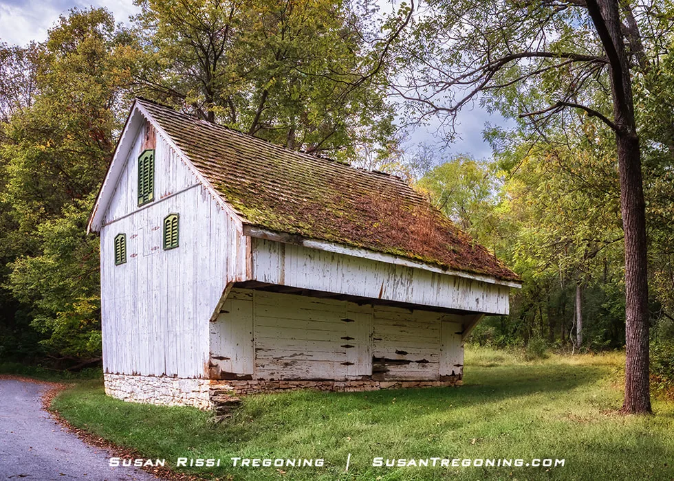 An old whitewashed wooden barn with a steep moss‑covered roof leans slightly beside a narrow paved path. The structure is surrounded by lush green grass and dense trees under soft, filtered daylight.