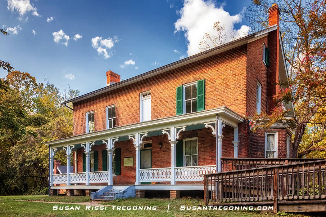 A two‑story historic brick house with green shutters and a white columned porch stands among early autumn trees, with a wooden accessibility ramp extending from the porch under a partly cloudy sky.