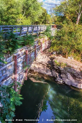 A historic stone aqueduct spans a calm, tree‑lined waterway, its metal supports and deck partially shaded by dense green foliage, with sunlight reflecting off the water below.