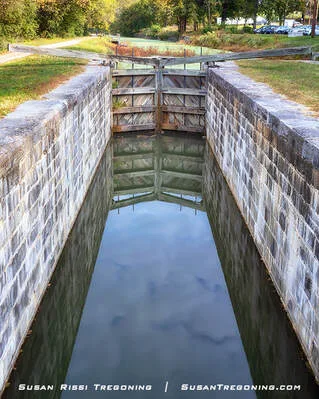 Stone‑lined canal lock filled with still water, reflecting trees and sky. Wooden lock gates stand at the far end, with grassy banks and scattered trees surrounding the lock.