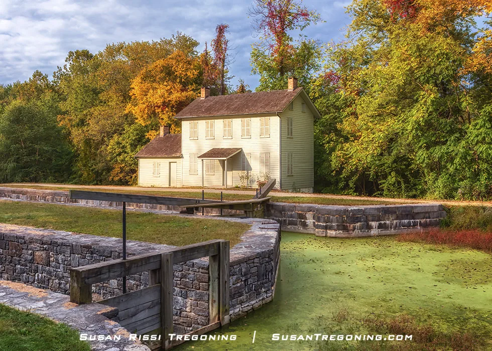 A two‑story light‑colored wooden lockhouse with a gabled roof stands beside a stone canal lock filled with green, algae‑covered water. Wooden lock gates frame the foreground, and dense autumn trees surround the scene, creating a quiet historic setting.