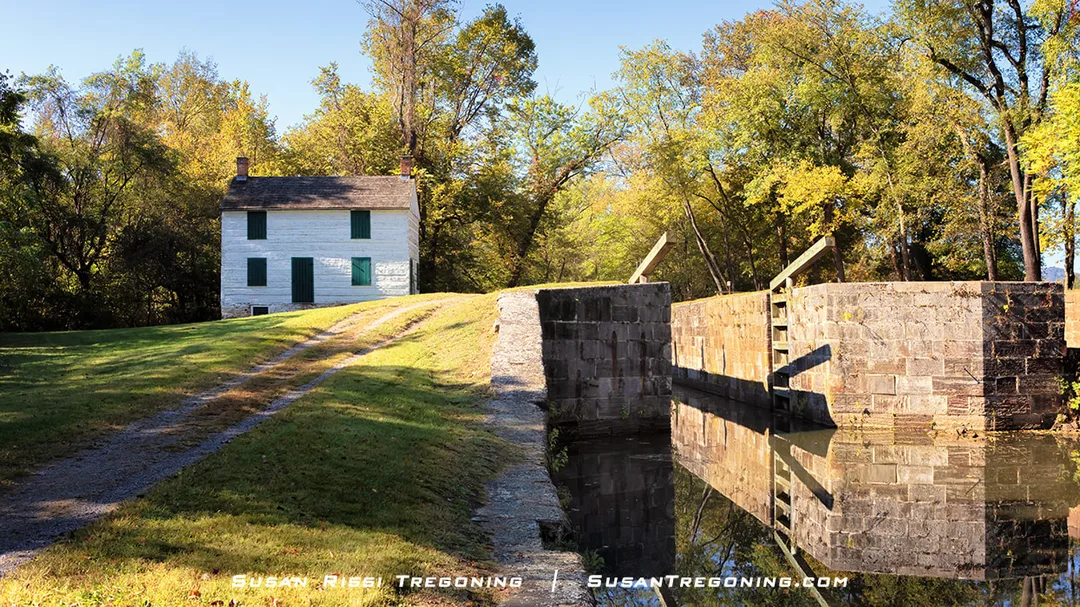 A white two‑story lockhouse with green shutters stands on a grassy slope beside a stone canal lock filled with still water, surrounded by early‑autumn trees and a gravel path leading toward the house.