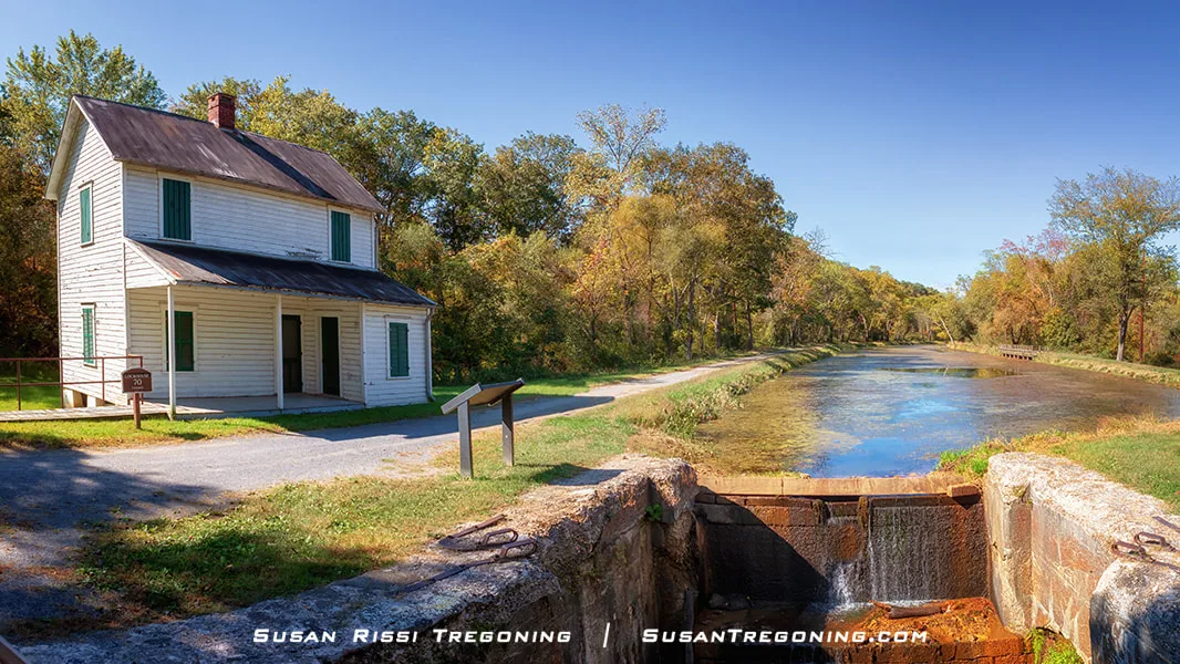 A white two‑story lockhouse with green shutters stands beside a stone canal lock filled with gently flowing water, surrounded by early autumn trees and a gravel path leading into the distance.
