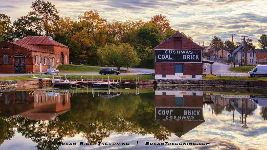 Historic brick buildings at Cushwa’s Warehouse reflect in the still canal water at Williamsport, Maryland. One building displays the “Cushwa’s Coal Brick” sign, and autumn trees line the background under a partly cloudy sky.