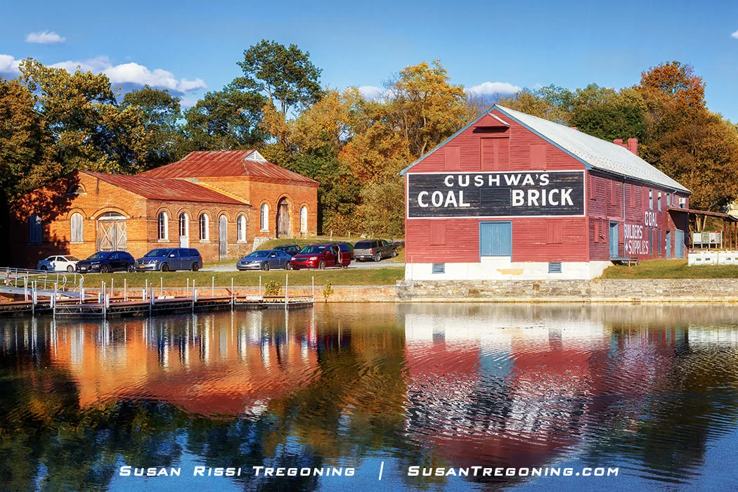 A pair of historic brick buildings stand beside the calm water of the Cushwa Turning Basin, their reflections visible on the surface. The larger red warehouse displays the painted “Cushwa’s Coal Brick” sign, while an older brick structure with arched windows sits nearby. Cars are parked between the buildings, and autumn trees fill the background.