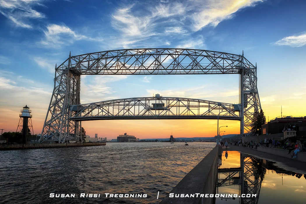 The Duluth Aerial Lift Bridge begins to rise at sunset, its steel structure reflecting warm orange and yellow light. A lighthouse stands to the left near the water’s edge, while people gather along the walkway on the right. The canal and sky are illuminated by the colors of the setting sun.