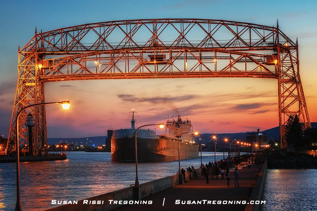 The H. Lee White departs Duluth at sunset, heading toward the Aerial Lift Bridge with warm light reflecting across the water. The bridge is partially raised, and people stand along the walkway watching the ship pass under a sky of orange, pink, and blue.