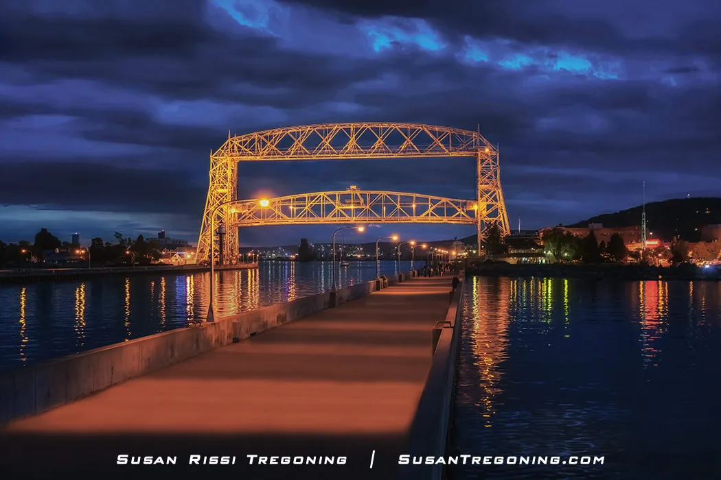 The Aerial Lift Bridge is illuminated at night, partially raised above the Duluth Ship Canal. Warm lights from the bridge and nearby waterfront buildings reflect across the dark water, and a walkway with streetlights leads toward the bridge under a cloudy evening sky.